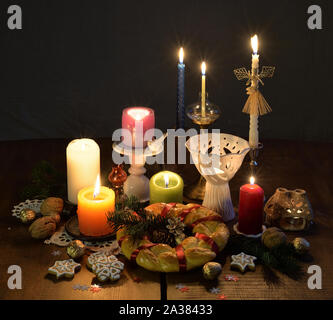 Burning candles in the candlesticks, a round ritual bread, gingerbread biscuits,  walnuts and straw angel figurine are on the wooden table in the dark Stock Photo