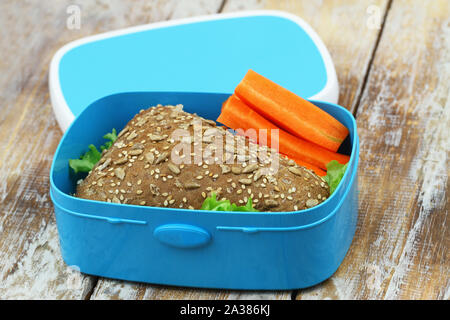 Brown sandwich with cheese and lettuce and crunchy slices of fresh carrots in blue lunch box on rustic woode surface Stock Photo