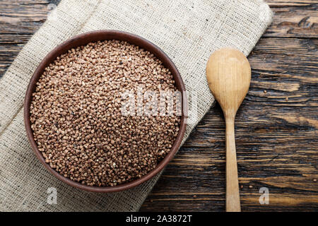 raw buckwheat in a clay plate, on a burlap napkin, on a wooden background. view from above. nearby lies a spoon. place for text Stock Photo