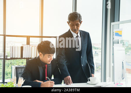 Senior businessman explaining business matters to the junior businessman. Adult manager teaches, suggests working for new businessmen at the desk. Stock Photo