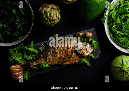 Sea bream baked in oven on black serving plate with arugula and green vegetables on black background. Flat lay, overhead shot. Stock Photo