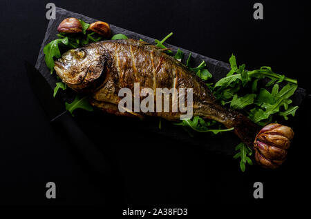 Baked dorada fish garnished with arugula on black background. Dark food photo. Seafood concept. Overhead shot. Copy space Stock Photo