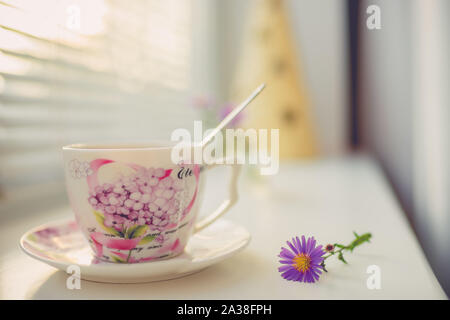 Elegant vintage porcelain cup and spoon on the white windowsill, side view with warm sunlight. Stock Photo