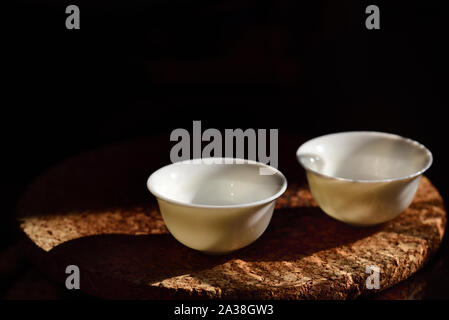 Close up of two empty small white chinese tea cups standing against a dark background on a cork plate with free space for text at a Chinese tea ceremo Stock Photo