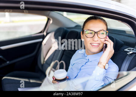 Attractive young woman on the business trip, taking a coffee break. Attractive business woman with phone in the back seat of the car. Close up portrai Stock Photo