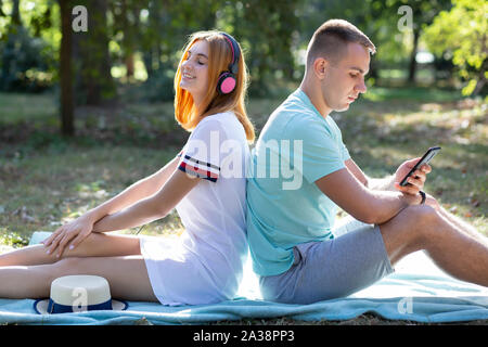 Young teenage couple having fun outdoors in summer park. Girl with red hair listening to music in pink earphones and boy chatting on the sell phone. Stock Photo