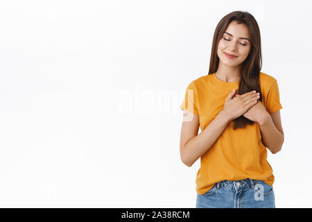 Tender, carefree romantic, dreamy young brunette woman in yellow t-shirt, holding hands pressed to heart, filled with warmth of love and passion Stock Photo