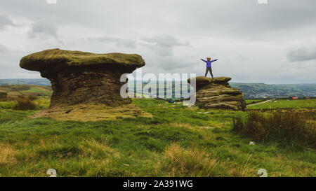 Person standing on top of the Doubler Stones with arms outstretched, Ilkley Moor, UK Their unusual shape is caused by the top being made of hard grits Stock Photo