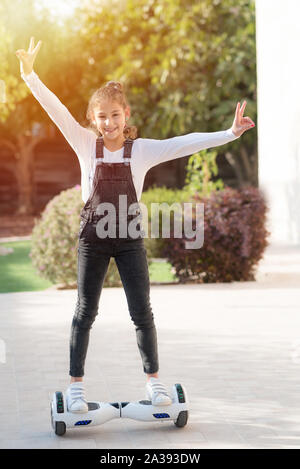 Young happy teenager girl with hands and victory sign up balancing on electric hoverboard at the sunny park, flare light.Child using a self-balancing two-wheeled board.Beautiful kid Smile with Braces Stock Photo