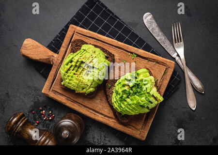 Vegan avocado rye bread toast with parsley and spices served on wooden cutting board over black concrete background. Table top view. Clean eating conc Stock Photo