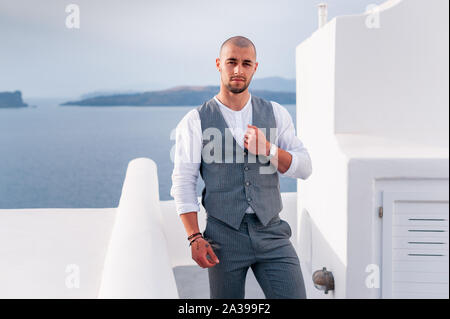 happy stylish groom smiling on background blue sea and sky in island Santorini. Young groom on the background of sea and architecture, Santorini island Greece. Stock Photo
