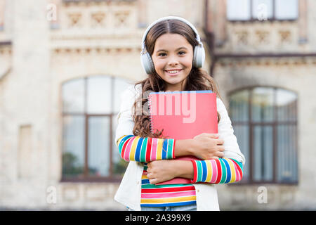 Audio programmes for pupils. Happy girl listening to audio book playing in headphones. Small child enjoying audio learning. Little kid in stereo earphones holding audio materials for lesson. Stock Photo