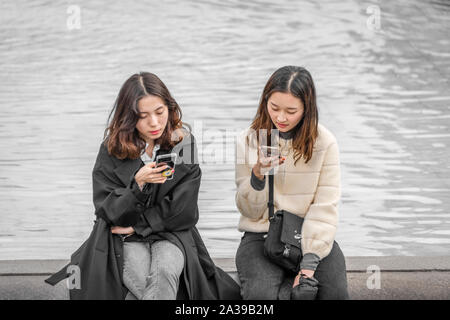 Two young girls check their phones whilst sitting on the edge of a fountain in Trafalgar Square, central London. Stock Photo