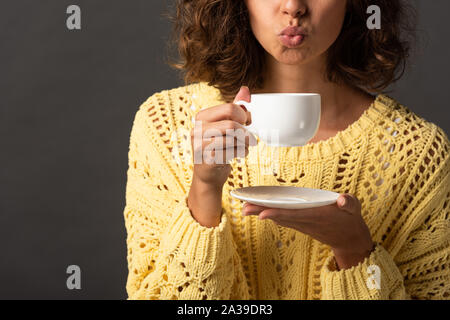 cropped view of curly woman in yellow knitted sweater pouting lips while holding cup of coffee and saucer on black background Stock Photo