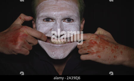 Clown Halloween man portrait. Close-up of an crazy, evil clowns face. White face makeup. Green hair. Scary smile with fingers. Attractive model in Halloween costume. Dark background Stock Photo