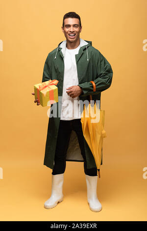 mixed race man in raincoat and rubber boots holding gift box and umbrella isolated on yellow Stock Photo