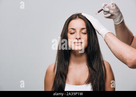 cosmetologist in latex gloves doing scalp mesotherapy to brunette beautiful woman with long hair and closed eyes isolated on grey Stock Photo