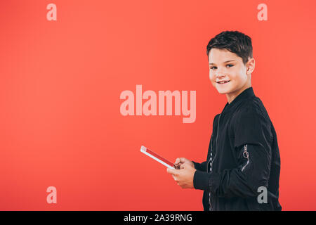 smiling kid in jacket using digital tablet isolated on red Stock Photo