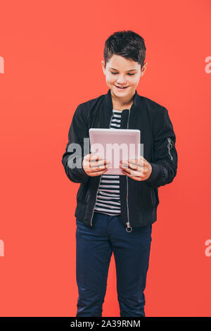 smiling kid in jacket using digital tablet isolated on red Stock Photo
