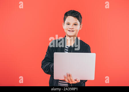 front view of smiling kid in jacket using laptop isolated on red Stock Photo