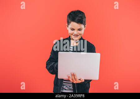 front view of smiling kid in jacket using laptop isolated on red Stock Photo