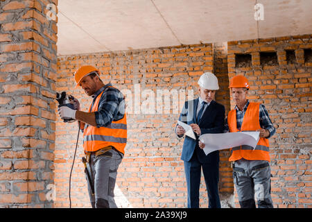 handsome builder using hammer drill near businessman and coworker Stock Photo