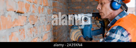 panoramic shot of mature builder using hammer drill near brick wall Stock Photo