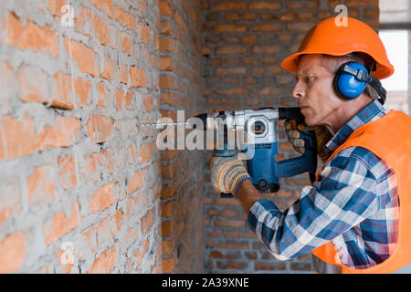 mature builder using hammer drill near brick wall Stock Photo