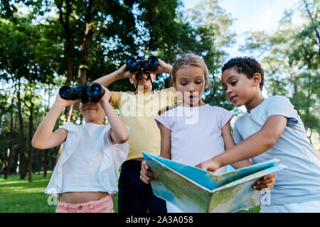 surprised kid looking at map with african american boy near friends with binoculars in park Stock Photo