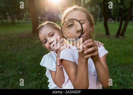 cheerful kid holding magnifier near face while standing with friend in park Stock Photo
