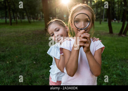 happy kid holding magnifier near face while standing with friend in park Stock Photo