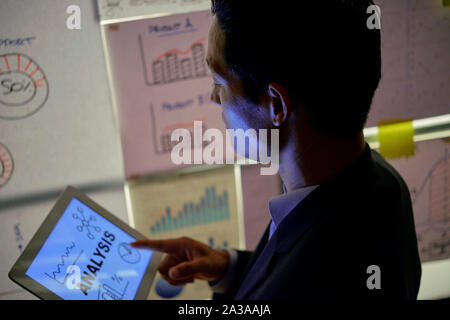 Serious Vietnamese entrepreneur standing at wall covered with paper reports and reading financial analysis on tablet computer Stock Photo