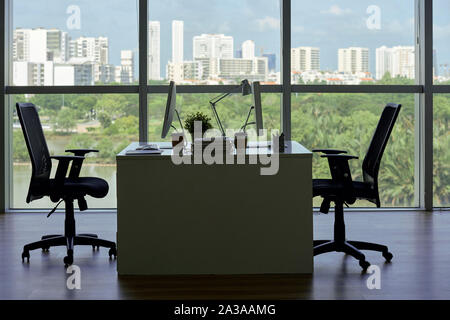 Desk with two chairs, computers and coffee cups in empty office, big panoramic window with view of big city in background Stock Photo