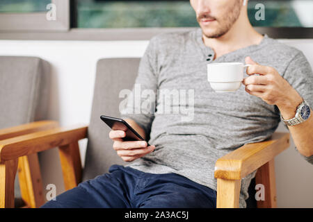 Cropped image of man resting in armchair, drinking cup of tea and reading news on smartphone Stock Photo