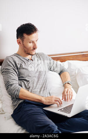 Handsome bearded young man sitting in bed and working on laptop Stock Photo