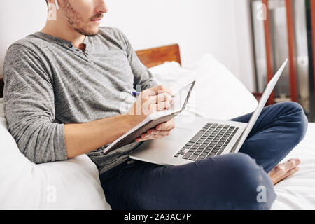 Cropped image of man wearing earbuds when watching webinar on laptop and taking notes in planner Stock Photo