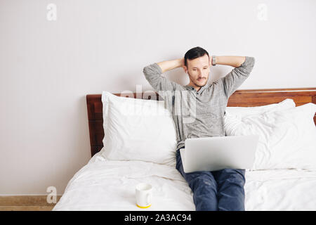 Handsome bearded man leaning back in his bed when watching new episode of popular show online Stock Photo