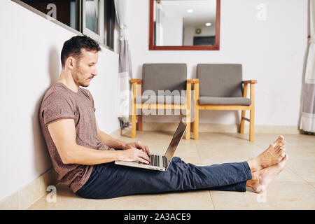 Barefoot young man sitting on the floor under opened window and programming on laptop Stock Photo