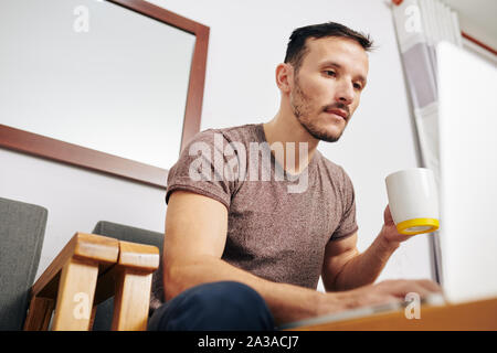 Handsome young man drinking big mug of coffee and reading article on laptop screen Stock Photo