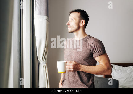 Handsome pensive young man drinking big mug of coffee and looking through window of his bedroom in the morning Stock Photo