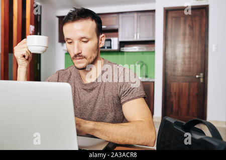 Bearden pensive man drinking cup of coffee and looking at programming code on laptop screen Stock Photo