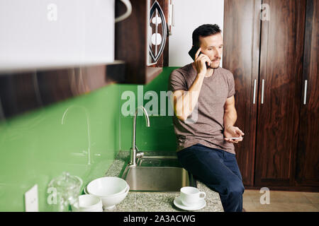 Handsome young man sitting on kitchen counter, talking on phone and ordering pizza and drinks Stock Photo