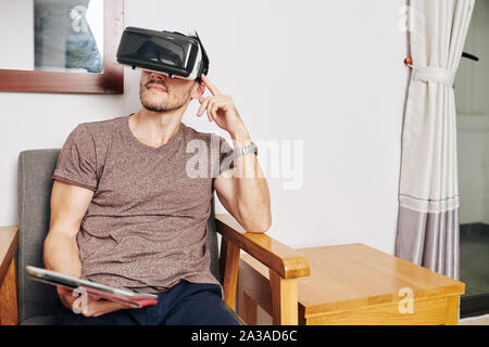 Man with tablet computer in hands using virtual reality headset when sitting on chair at home Stock Photo