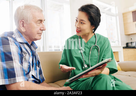 Smiling female hospital worker visiting senior patient at home, showing him information on tablet computer and discussing medical tests results Stock Photo