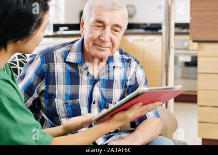 Smiling senior man reading information on tablet computer in hands of nurse who is visiting him at home Stock Photo