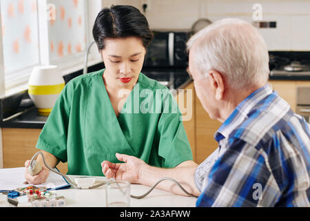 Serious young Asian medical nurse using tonometer to measure blood pressure of senior patient when visiting him at home Stock Photo