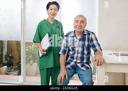 Portrait of smiling senior man sitting next to his medical nurse visiting him at home for weekly check-up Stock Photo