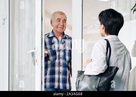 Smiling senior man in plaid shirt opening glass door to invite caregiver to house Stock Photo