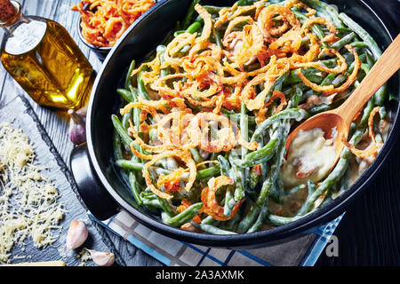 Homemade Green Bean Casserole topped with French crispy fried onions in a black dish with spoon on a wooden table,  american cuisine, close-up Stock Photo