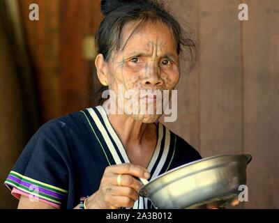 Middle-aged Chin Muun tribal woman ('spider woman') with traditional facial tattoo eats from a stainless-steel bowl and looks at the camera. Stock Photo